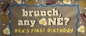 Hand-painted kraft paper banner reading "brunch, any one? Ben's first birthday", with fried eggs, bacon, and waffles painted around a blue scalloped border.